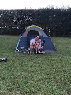 [IMAGE DESCRIPTION: A photo of a tent pitched in a backyard. An adult with short hair and a child sit outside the tent, smiling at the camera.]