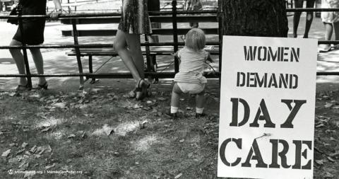 Black and white image of the back of child and people walking by next to a sign that reads "Women Demand Day Care"
