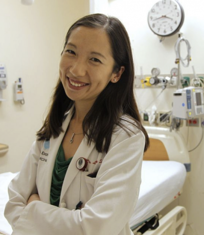 Person with long hair, wearing a white doctor's coat, smiles at the camera.