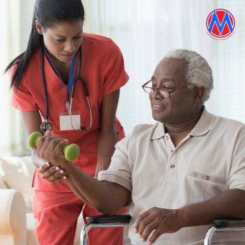 A nurse wearing red scrubs assists an elderly patient in a wheelchair lift a weight with their right arm. 