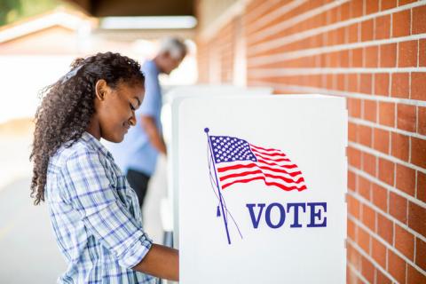 [IMAGE DESCRIPTION: Person with long dark curly hair stands at a voting booth.]