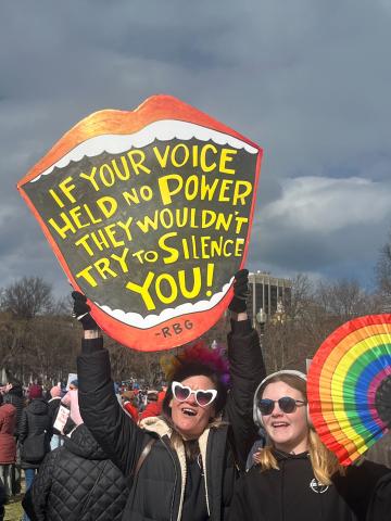 Our voice is our power — Join MomsRising in taking 5 urgent steps for our democracy after the historic 'No Kings' march. Two people at the No Kings march hold a mouth-shaped sign with an RBG quote and a rainbow fan.