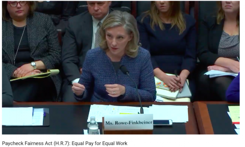 [IMAGE DESCRIPTION: A person with shoulder length blond hair and a blue jacket sits before a House committee, providing witness testimony on the need for equal pay for equal work.]