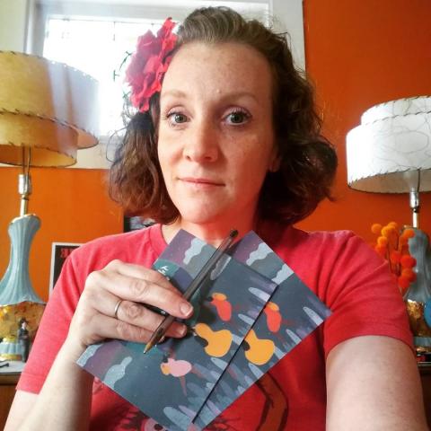 [IMAGE DESCRIPTION: A photo of a woman wearing a red flower in her chin length wavy brown hair, looking at the camera and holding up her get-out-the-vote postcards.]