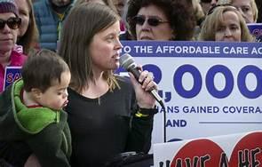 [IMAGE DESCRIPTION: A photo of a woman with shoulder length brown hair holding a microphone in one hand and a toddler in the other.]