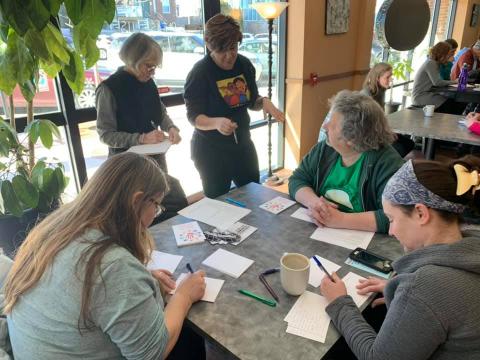 [IMAGE DESCRIPTION: A group of people gather around a table, writing postcards.]