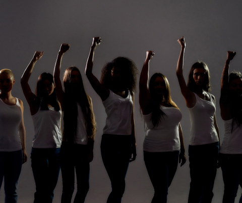 Variety of women standing side by side with their right fists raised in the air. 