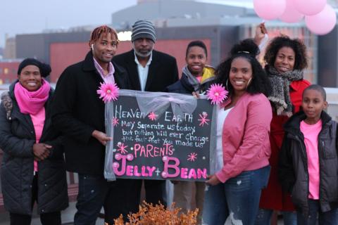 [IMAGE DESCRIPTION: A photo of seven people, adults and children, wearing black and bright pink. The two adults in the middle hold a sign to honor Jelly Bean.]