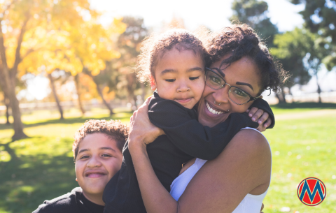 A mom with brown skin holds her toddler while her young son hugs her