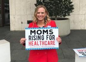 A blonde woman holds a sign that says Moms Rising for Health Care