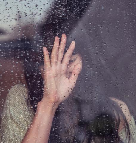 Woman at a rain covered window with her hand in front of her face.