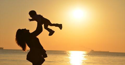 Woman holds her baby up in the air as the sun sets behind them on the beach
