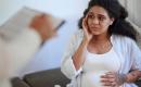 A concerned pregnant woman sitting on a couch, looking at a person holding a clipboard.