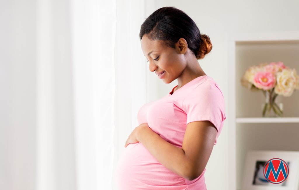 A woman with black hair and brown skin, wearing a pink shirt, cradles her pregnant belly