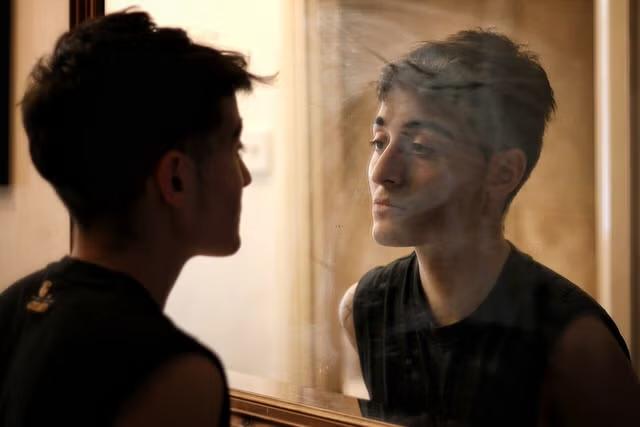 A person with short dark hair and medium skin contemplates their reflection in a mirror