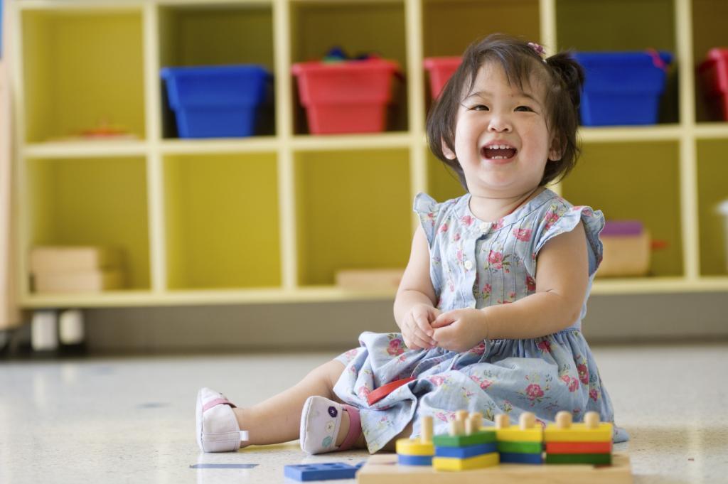 [IMAGE DESCRIPTION: A young child with shoulder length black hair wears a light blue dress, sits on the floor with bright color blocks, and laughs.] 