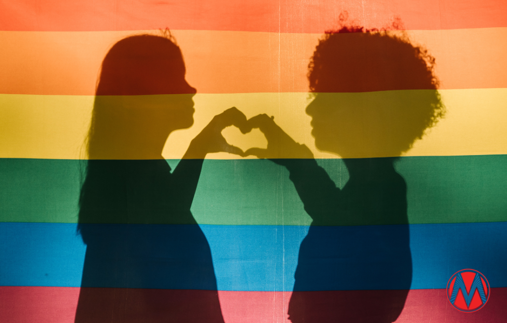 Two feminine-presenting silhouettes forming a heart with their hands behind a Pride flag
