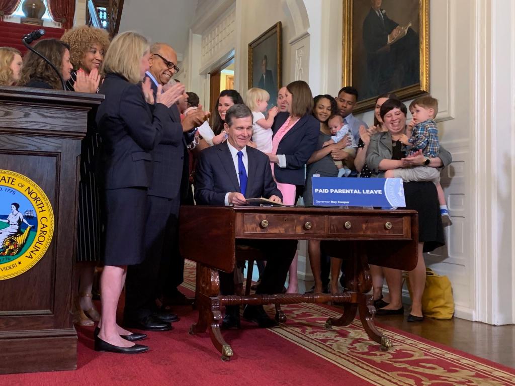 [IMAGE DESCRIPTION: The Governor of North Carolina sits behind a desk and smiles while signing paid leave into law. He is surrounded by a group of adults and some children, also smiling.]