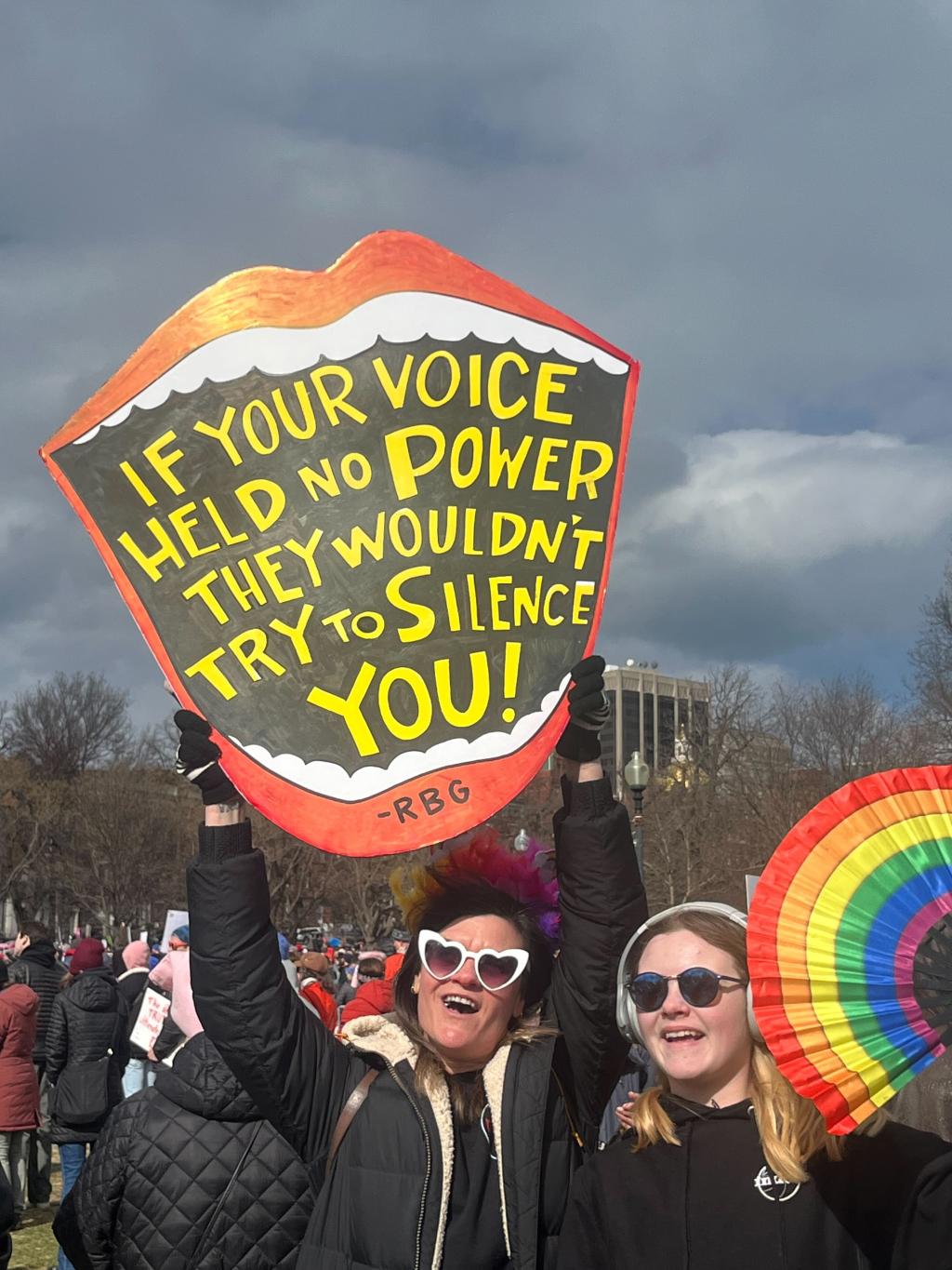 Our voice is our power — Join MomsRising in taking 5 urgent steps for our democracy after the historic 'No Kings' march. Two people at the No Kings march hold a mouth-shaped sign with an RBG quote and a rainbow fan.