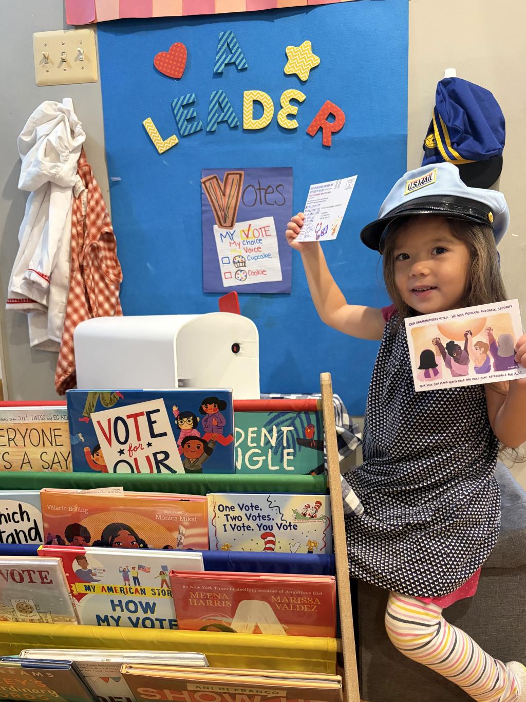Child holding MomsRising postcard around children's books and a sign titled "A Leader Votes"