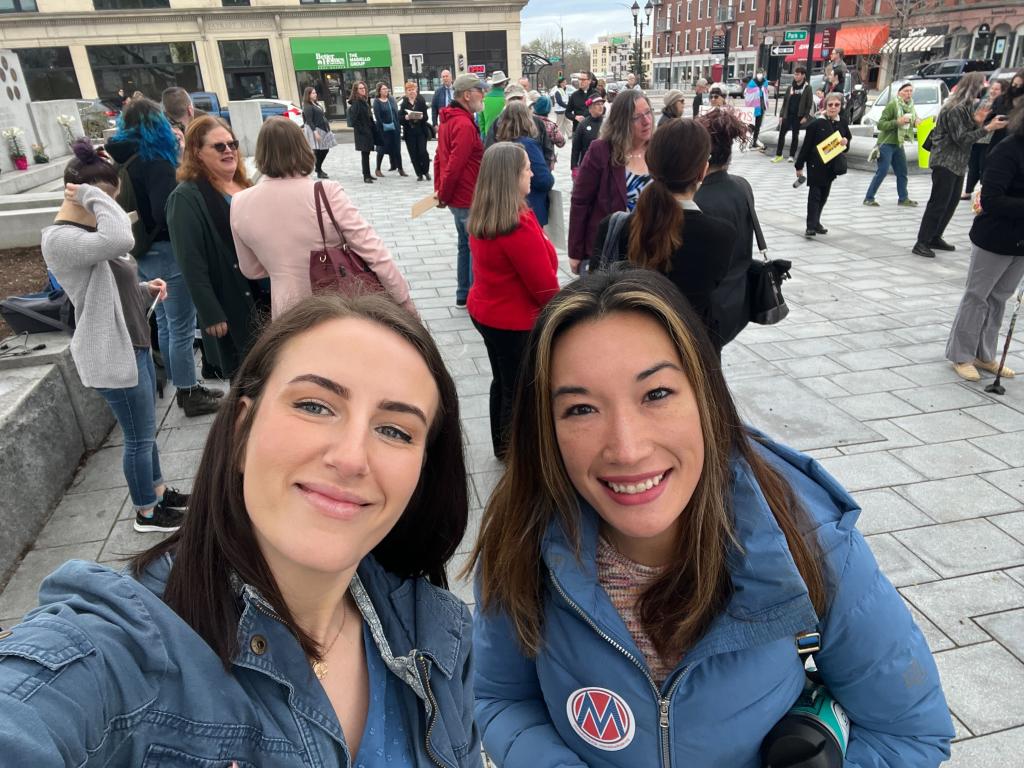 A white women and an Asian woman smiling at the camera with a crowd behind them