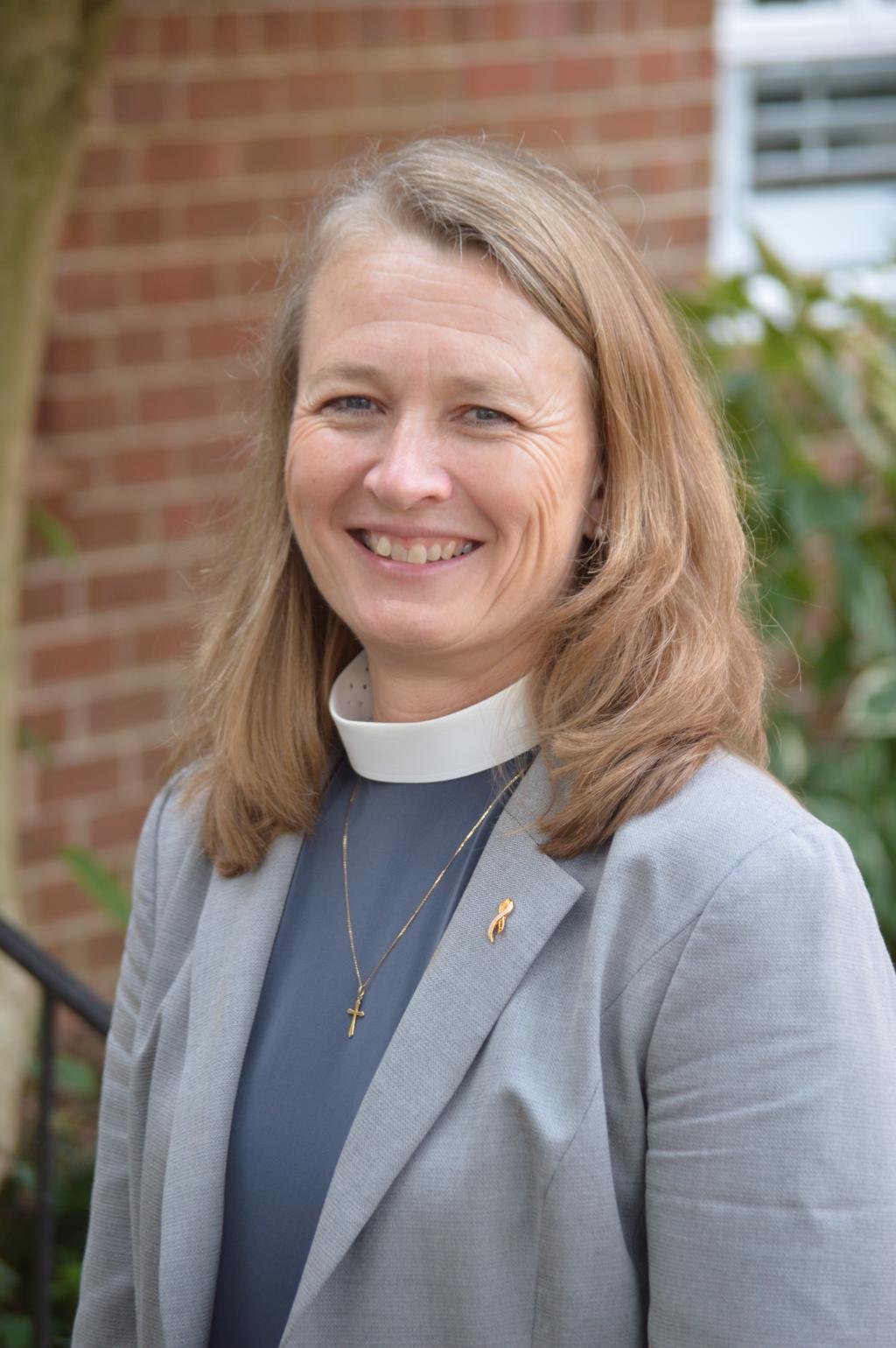 [IMAGE DESCRIPTION: A person with shoulder length strawberry blond hair and blue eyes smiles at the camera, wearing a clergy collar and blue shirt and gold cross.]