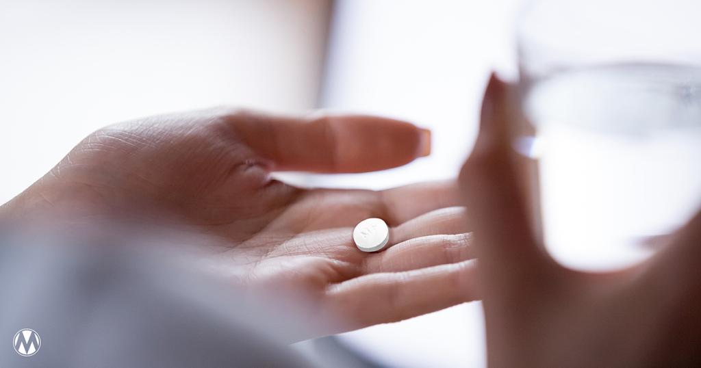 Close up of a hand holding a white mifepristone pill (labeled 'M') with a glass of water nearby. 