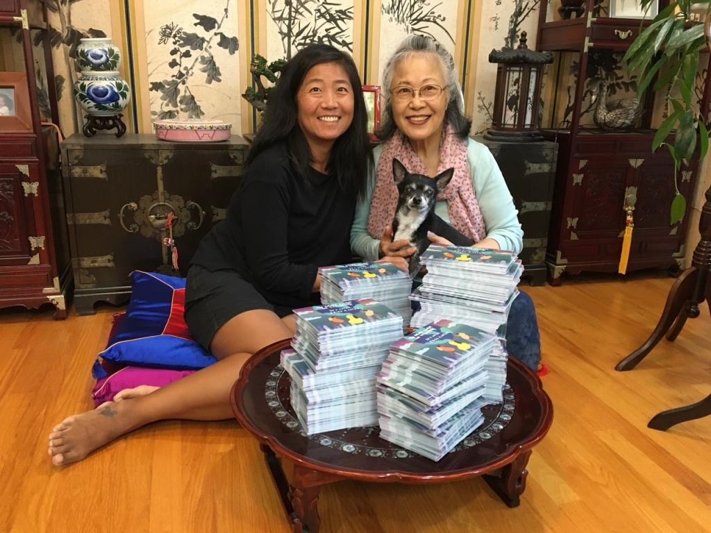 [IMAGE DESCRIPTION: A photo of a woman and her mother, seated on the floor in front of a low round table filled with 2500 handwritten postcards. One woman is holding a dog.]