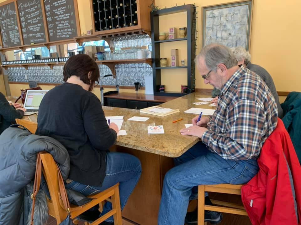 [IMAGE DESCRIPTION: A group of people gather at a cafe, writing postcards.]