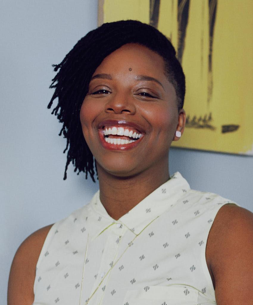 [IMAGE DESCRIPTION: A person smiles brightly at the camera, with shoulder length hair parted to the side, wearing a sleeveless shirt.]