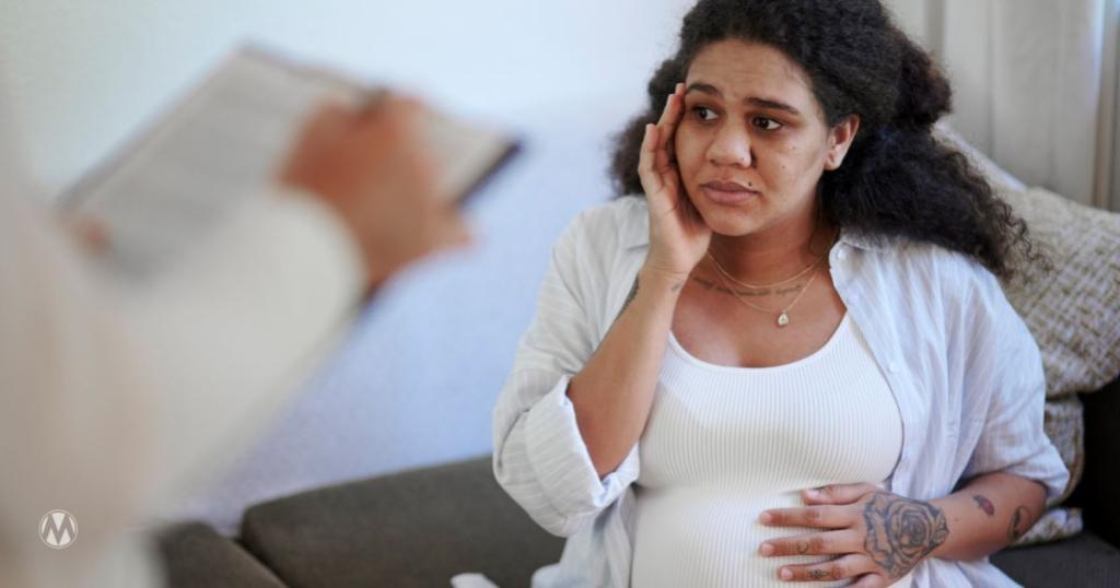 A concerned pregnant woman sitting on a couch, looking at a person holding a clipboard.