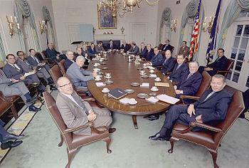 About 20 white men and 1 Black man in suits around a long table in the Whitehouse