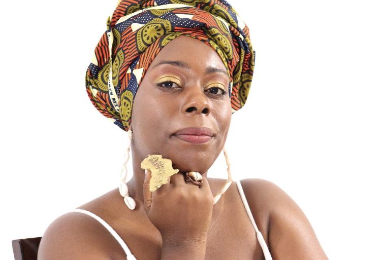 [IMAGE DESCRIPTION: A photo of a person looking at the camera with a slight smile, wearing a colorful headwrap and a matte gold ring in the shape of the African continent.]