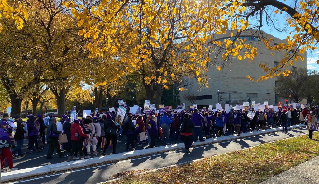 People marching with signs along Washington DC street and trees with changing leaves around them. 