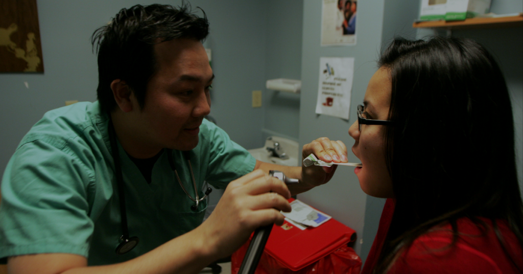 [IMAGE DESCRIPTION: In a medical clinic. A health professional in greens scrubs with short black hair peers inside the mouth of a patient with shoulder length black hair.]