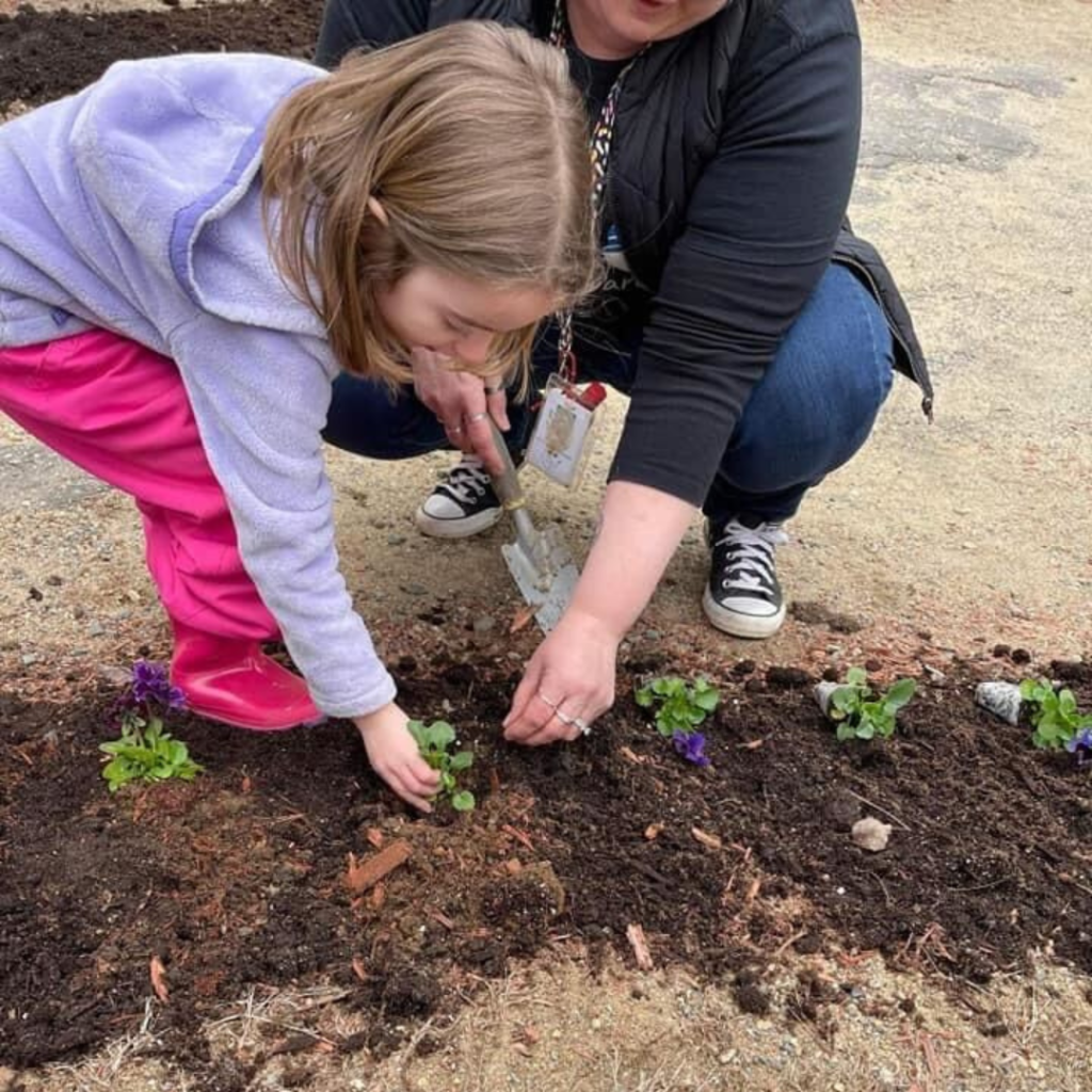 Little girl in purple jacket, pink pants and rain boots bending down to plant flowers with a teacher squatting down next to her.