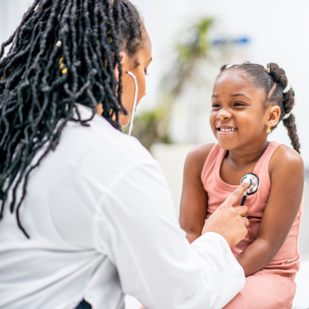 Black female doctor holds up a stethoscope to a smiling black female child.