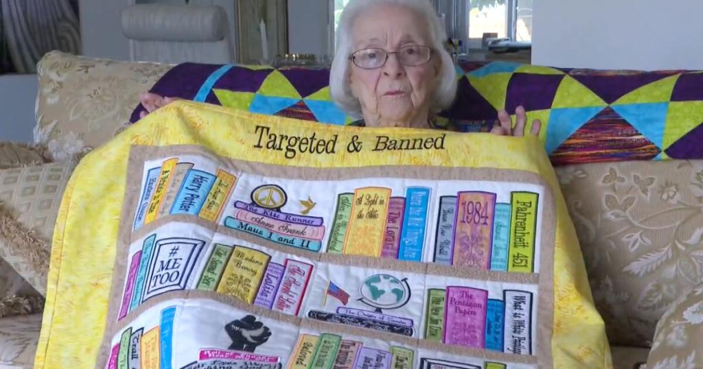 An elderly white woman holding up a quilt that looks like a bookshelf, which says "Targeted & Banned" stitched at the top