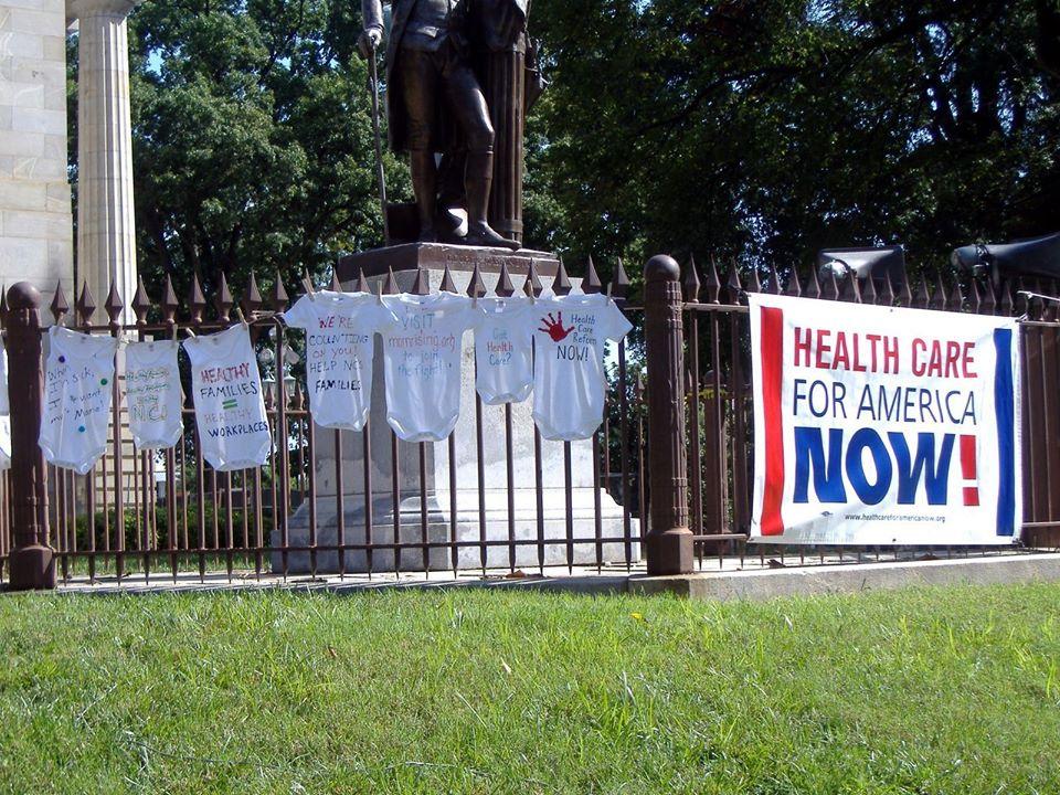 [IMAGE DESCRIPTION: A photo of baby shirts hung on a fence with pro-healthcare messages written on them.]