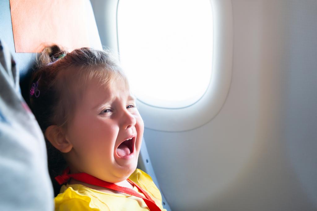 [IMAGE DESCRIPTION: A photo of a young child with blond hair in a half ponytail crying in an airplane seat.]