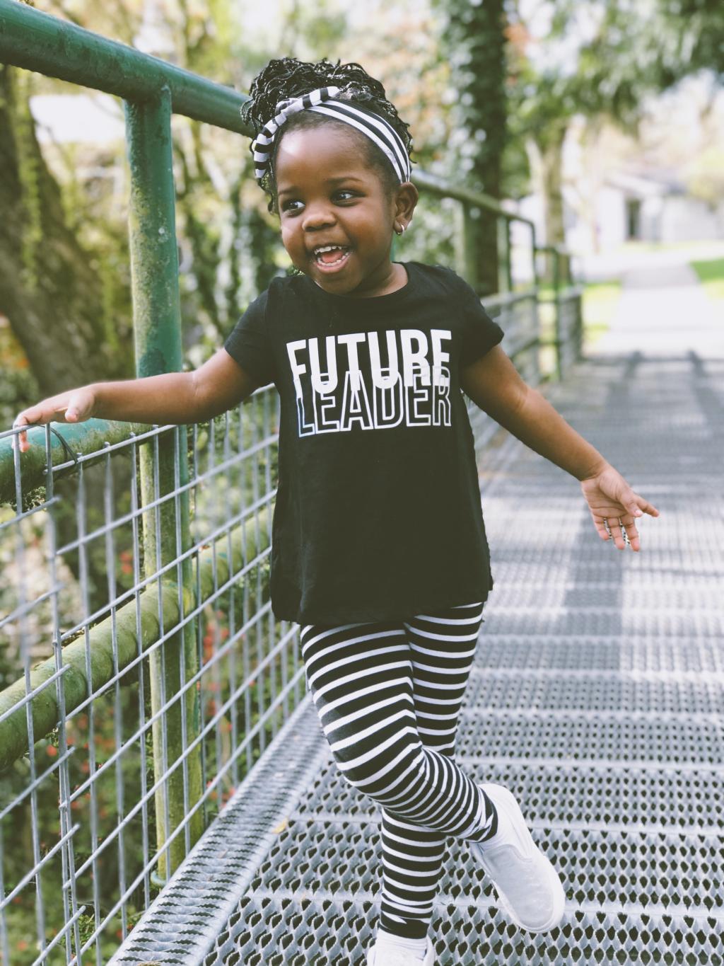 Little girl holding a handrail wearing striped pants, matching head wrap and shirt that reads "Future Leader"