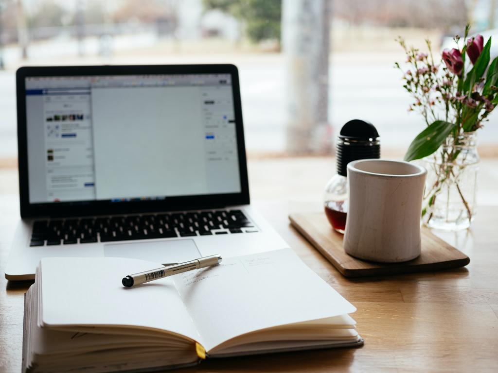 [IMAGE DESCRIPTION: A photo of a laptop on a desk with an open journal and pen in front of it, a coffee mug out of focus behind it.]
