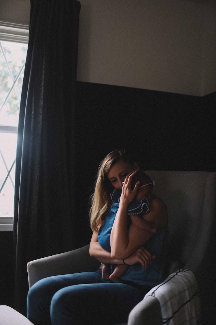 Photo by Jenna Norman on Unsplash. Mom holding baby in arms while seated. Sunlight is streaming in through a side window.