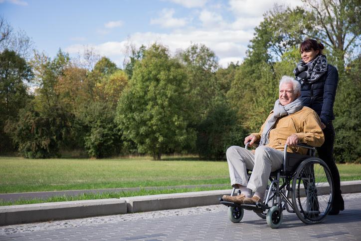 Person with white hair sits in wheelchair pushed by another person outdoors under a blue sky.