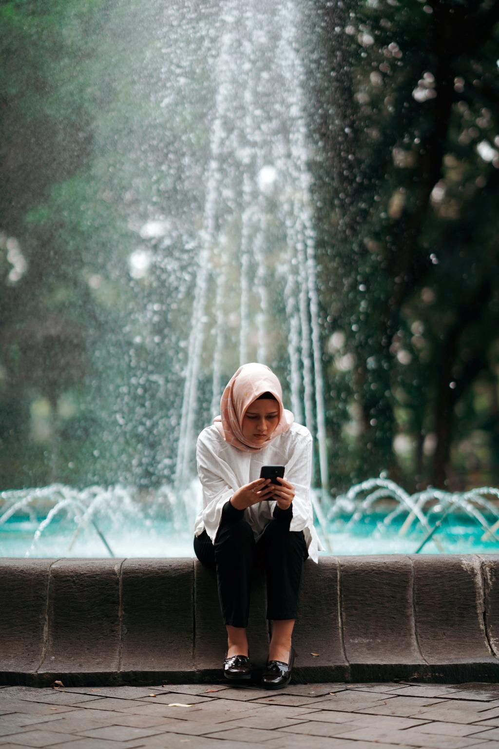 Photo of a woman sitting at an outdoor fountain, reading her phone