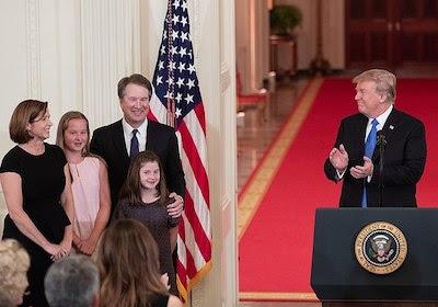 Judge Kavanaugh and family stand to the right of a podium, behind which Pres. Trump stands in mid clap looking at Kavanuagh