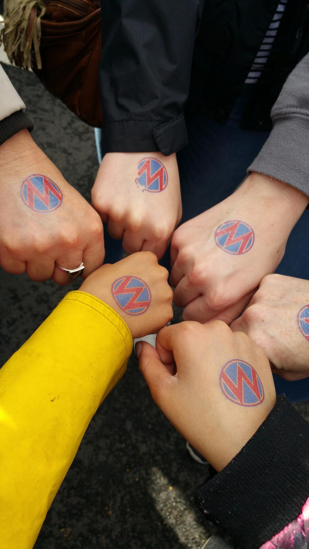 [IMAGE DESCRIPTION: A photograph of a circle of fists, kid-sized to adult, with MomsRising circle M tattoos.]