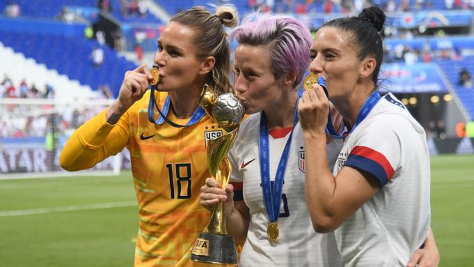 [IMAGE DESCRIPTION: Three players from the US Women's National Team for soccer smooch their medals and trophy.]